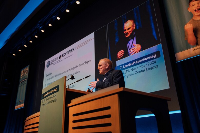Prof. Andreas Beelmann referiert im großen Saal vor Publikum hinter einem Rednerpult, hinter ihm ist seine Bildschirmpräsentation