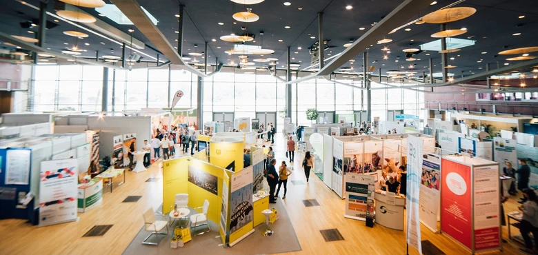 die Übersicht von einer Messe in einem großen Raum im Congress Center Dresden. Viele Ausstellende mit ihren Ständen an den vereinzelt Menschen stehen.
