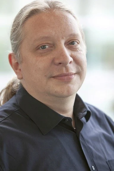 Das Bild zeigt ein Porträt von Sven Kruppik in einem schwarzen Hemd mit langen grauen Haaren zu einem Zopf gebunden, vor einem verschwommenen Hintergrund.