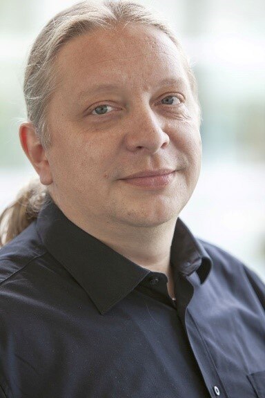 Das Bild zeigt ein Porträt von Sven Kruppik in einem schwarzen Hemd mit langen grauen Haaren zu einem Zopf gebunden, vor einem verschwommenen Hintergrund.