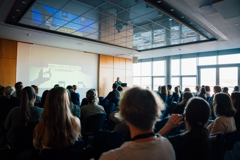 Menschen sitzen auf Stühlen und schauen nach vor zur Vortragenden Julia von Weiler. Neben ihr die Präsentation mit den Worten Analoge und Digitale Lebenswelt