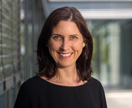 Portraitfoto von Verena Schreiber mit dunkelfarbenen Pulli links im Bild und im Hintergrund eine Fensterfront