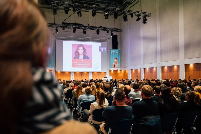 Professor Gerd Gigerenzer während seiner Livezuschaltung in den Saal 2, Menschen sitzen auf Stühlen in Reihen und schauen zur Bühne,