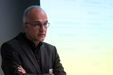Ein Mann mit verschränkten Armen schaut in den Raum, hinter ihm leicht verschwommen die Bildschirmpräsentation.am LPT6