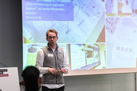 Ein junger Mann mit Brille, steht vor einer Bildschirmpräsentation und hält eine Vortrag.