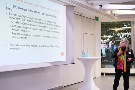 Eine Frau steht neben einem Stehtisch, sie hält ein Mikrofon in der Hand und schaut zu ihrer Bildschirmpräsentation.m LPT6 vor der Leinwand
