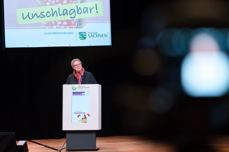 Eine Referentin hinter einem Rednerpult spricht in den Raum und hinter ihr ein Ausschnitt von einem Banner des LPT6. Zu sehen ist noch unschlagbar!.