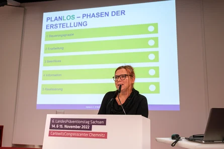 eine Frau an einem Rednerpult spricht in ein Mikrofon, hinter ihr ist ihre Bildschirmpräsentation.erpult