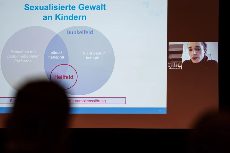 Eine Frau spricht in einem Onlinebeitrag. Sie ist mit einem kleinen Bild rechts neben der Bildschirmpräsentation. Vor der Präsentation sind die Schatten der Zuhörenden.