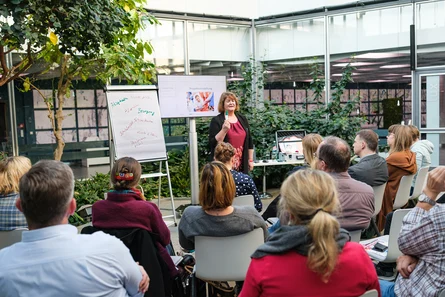 Eine Frau steht an einem Tisch hinter ihr ein Bildschirm und ein Flipchart und vor ihr sitzen mehrere Personen auf Stühlen.rkshopraum Lux beim Vortrag mit Publikum